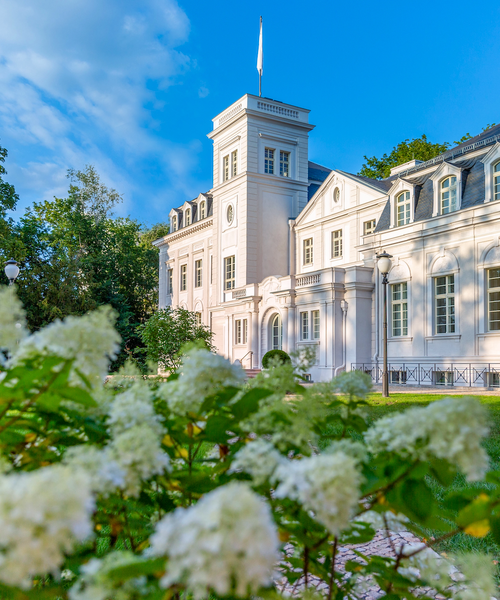 Helles historisches Gebäude mit Turm und weißen Fassadendetails, umgeben von blühenden Sträuchern und Grünflächen.