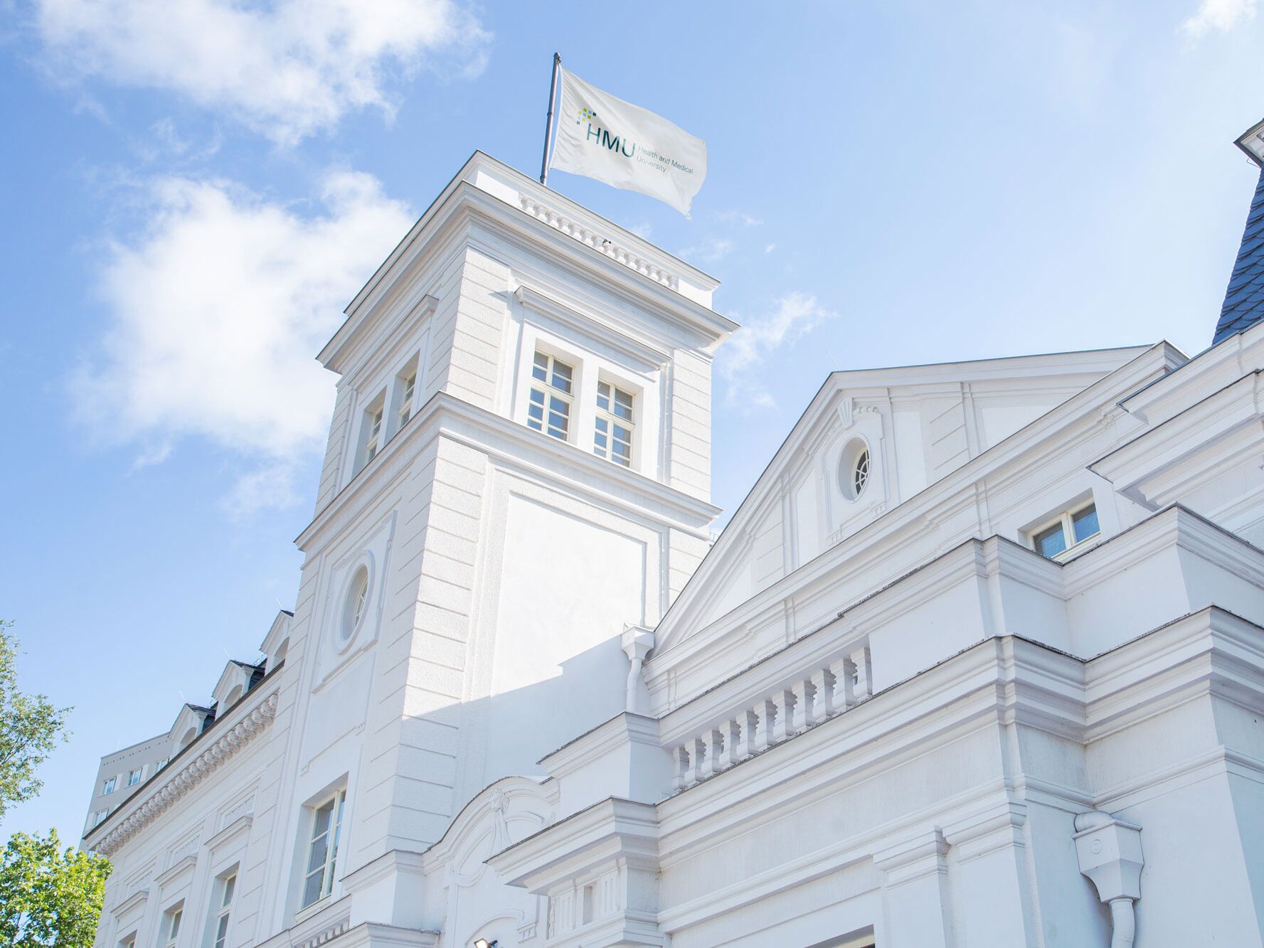 Weißes historisches Gebäude mit Turm und wehender HMU-Flagge vor blauem Himmel.