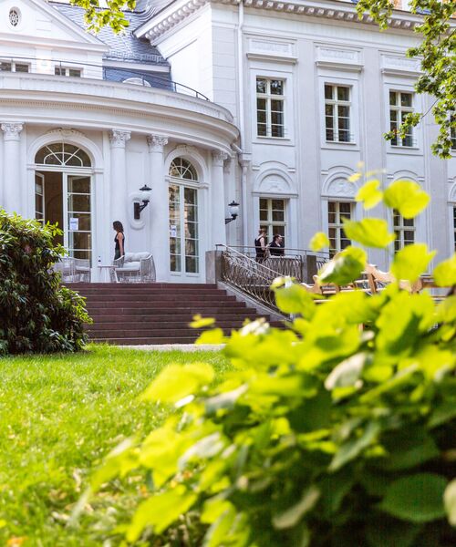 Blick auf die Terrasse eines eleganten weißen Gebäudes mit großen Fenstern, umgeben von Garten und Grünpflanzen.