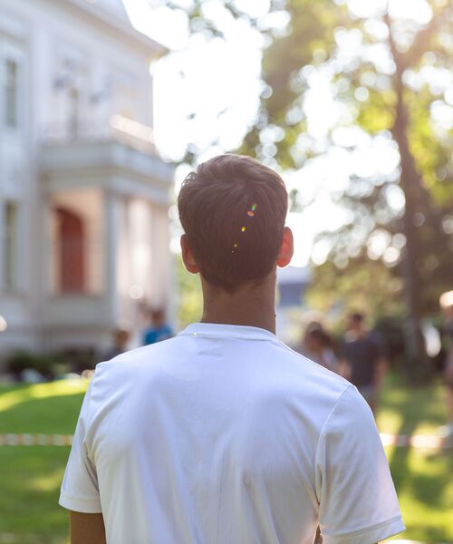 Person steht mit dem Rücken zur Kamera auf einer sonnigen Grünfläche vor einem hellen Gebäude.
