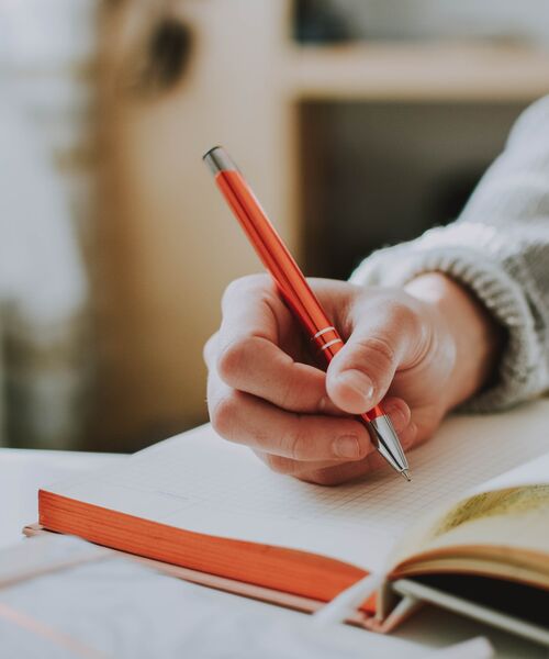 Hand schreibt mit einem roten Stift in ein geöffnetes Notizbuch.