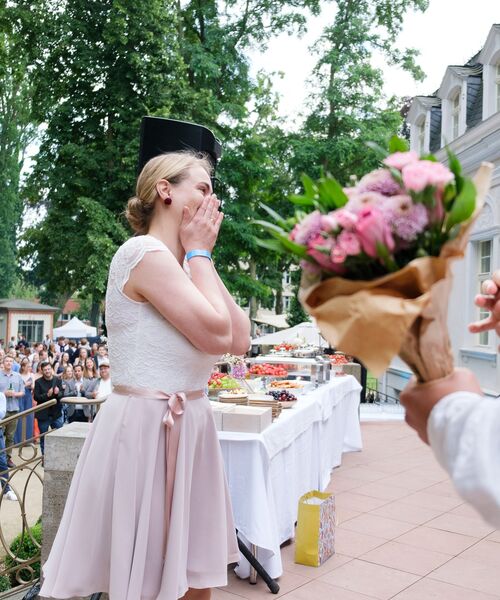 Junge Frau in festlicher Kleidung reagiert überrascht, während ihr ein Blumenstrauß überreicht wird.