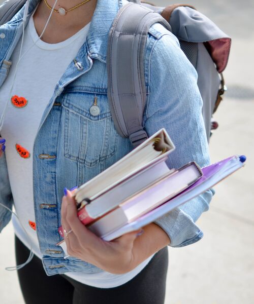 Eine Person mit Rucksack trägt mehrere Bücher und Hefte eng an den Körper gedrückt.