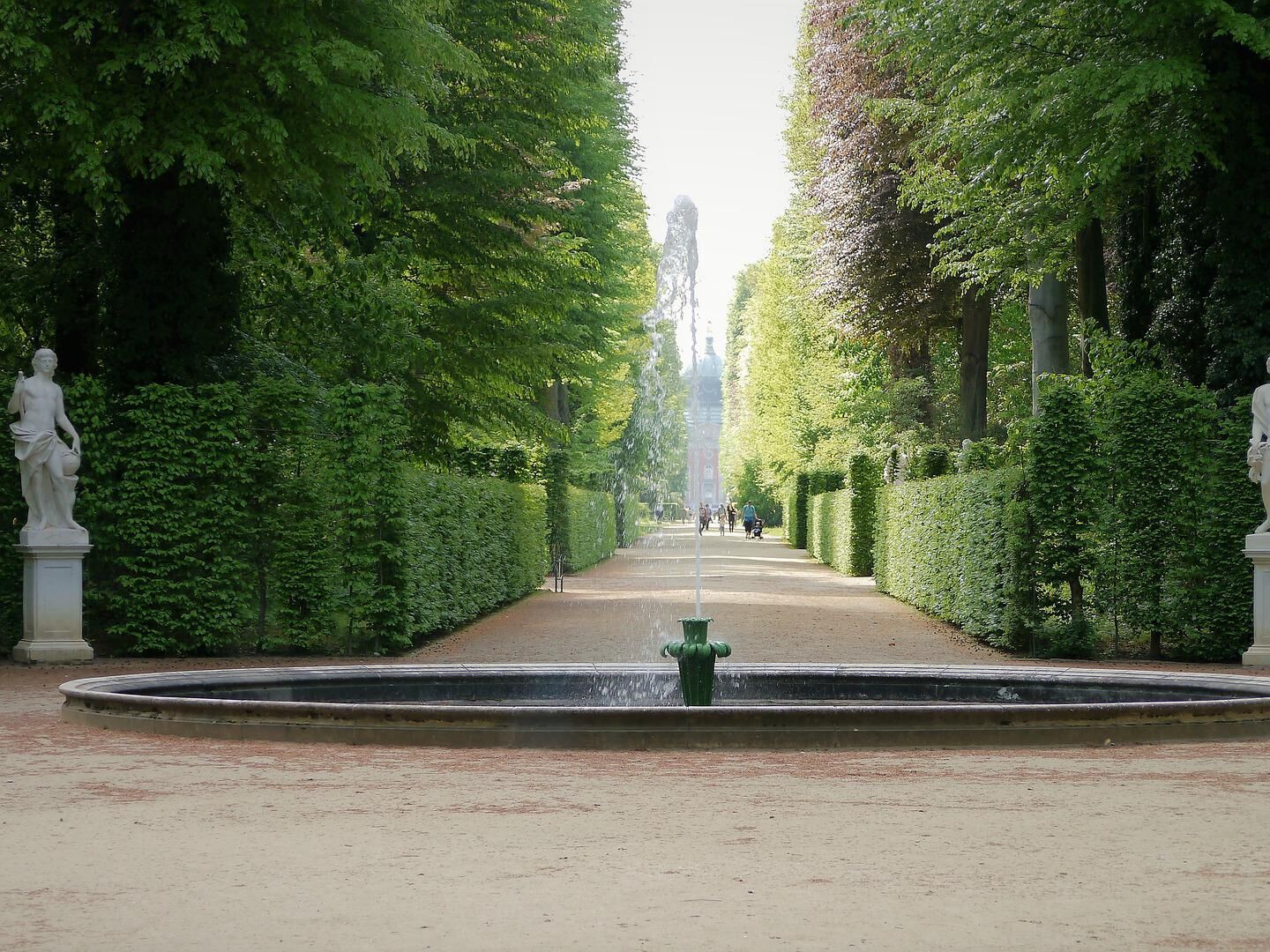 Geradlinige Gartenallee mit Springbrunnen, flankiert von hohen, dicht geschnittenen Hecken.