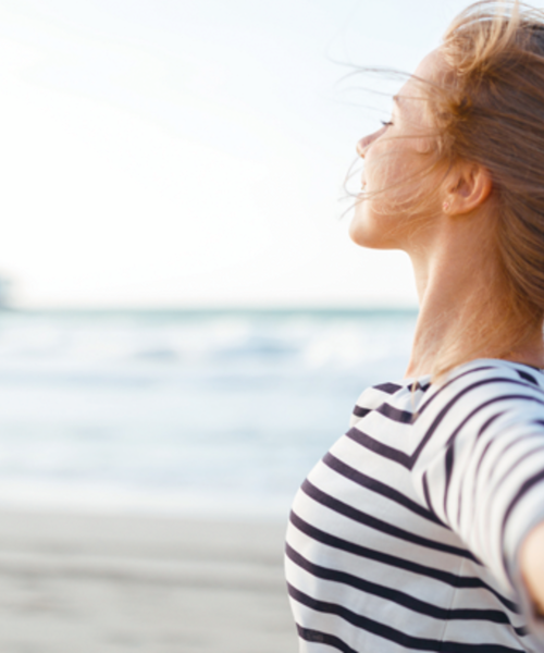 Frau steht am Strand, blickt erleichtert zur Seite und breitet die Arme aus.