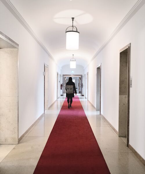 Person geht durch einen hellen Flur mit rotem Teppich und modernen Lampen, Blickrichtung zum Ausgang.