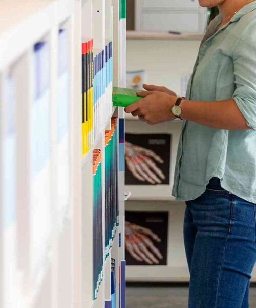 Person greift in einem Regal mit farblich sortierten Büchern und Fachliteratur nach einem Band.