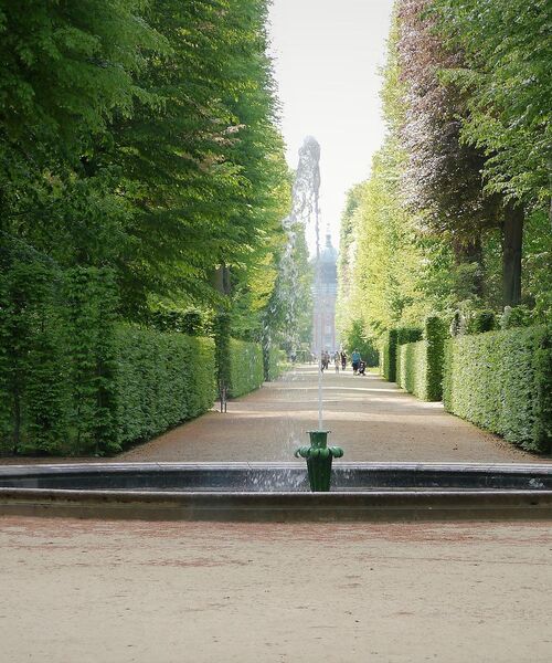 Geradlinige Gartenallee mit Springbrunnen, flankiert von hohen, dicht geschnittenen Hecken.