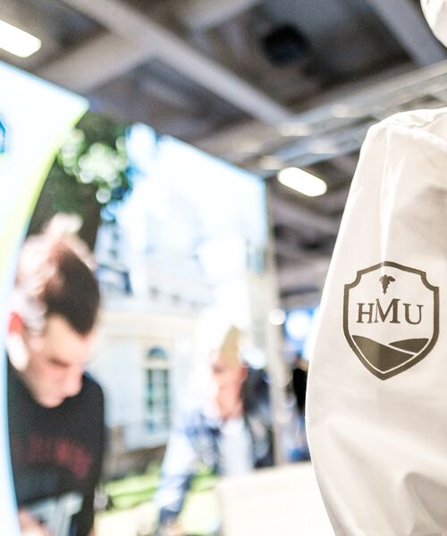 Detail einer Jacke mit HMU-Logo im Vordergrund, im Hintergrund unscharfe Personen und Messestand.
