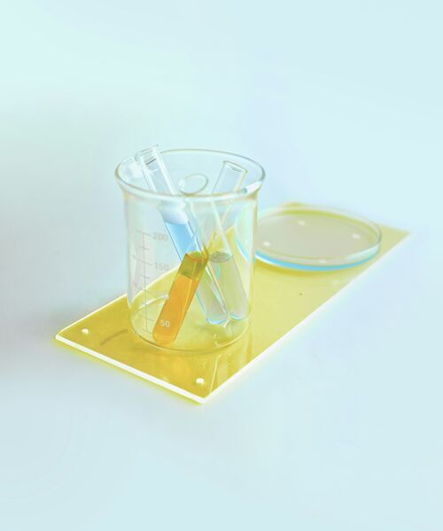 Messbecher mit mehreren Reagenzgläsern in unterschiedlichen Farben, daneben eine Petrischale auf heller Fläche.