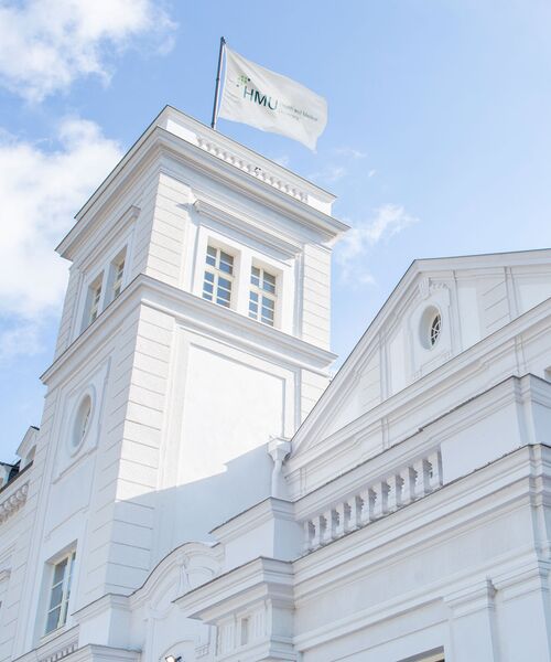 Weißes historisches Gebäude mit Turm und wehender HMU-Flagge vor blauem Himmel.