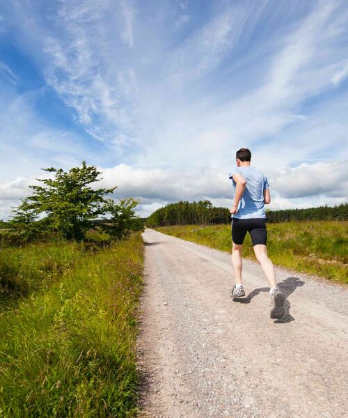 Person joggt auf einem langen, unbefestigten Weg durch eine offene Landschaft unter blauem Himmel.