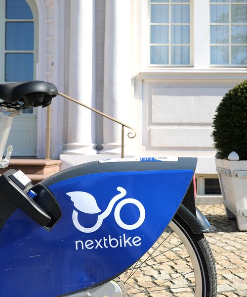 Blaues Nextbike-Leihfahrrad vor der hellen Fassade eines Gebäudes mit großer Fensterfront.