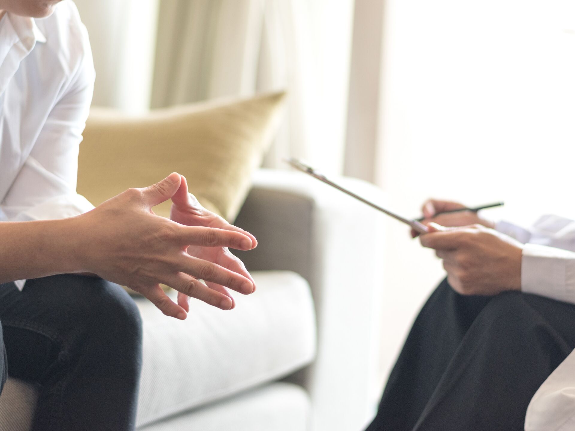 Zwei Personen im Gespräch auf einem Sofa, eine Person spricht mit gestikulierenden Händen, die andere hält einen Schreibblock.