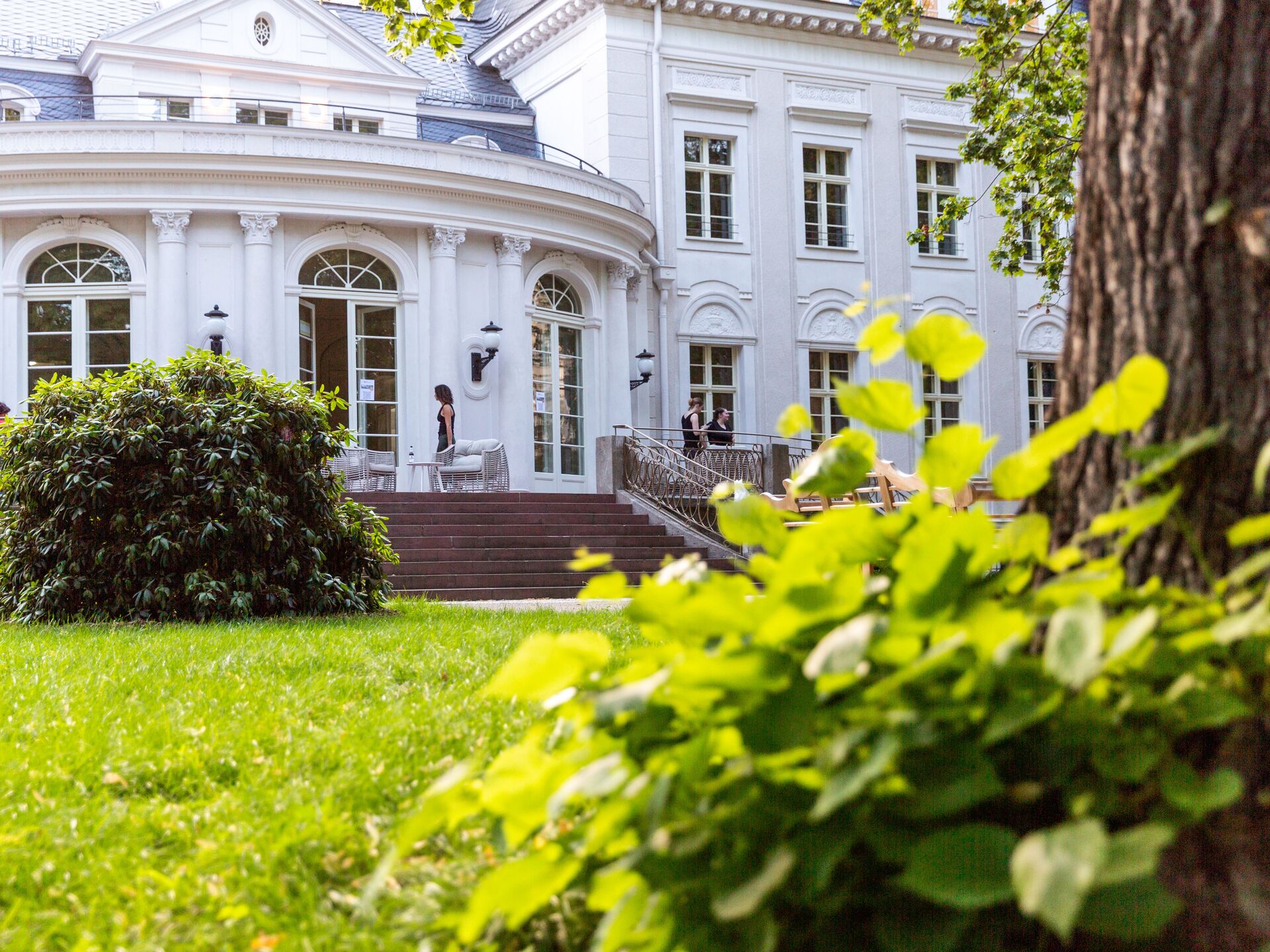 Blick auf die Terrasse eines eleganten weißen Gebäudes mit großen Fenstern, umgeben von Garten und Grünpflanzen.