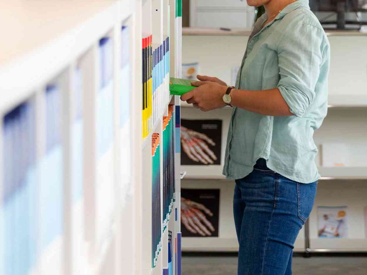 Person greift in einem Regal mit farblich sortierten Büchern und Fachliteratur nach einem Band.