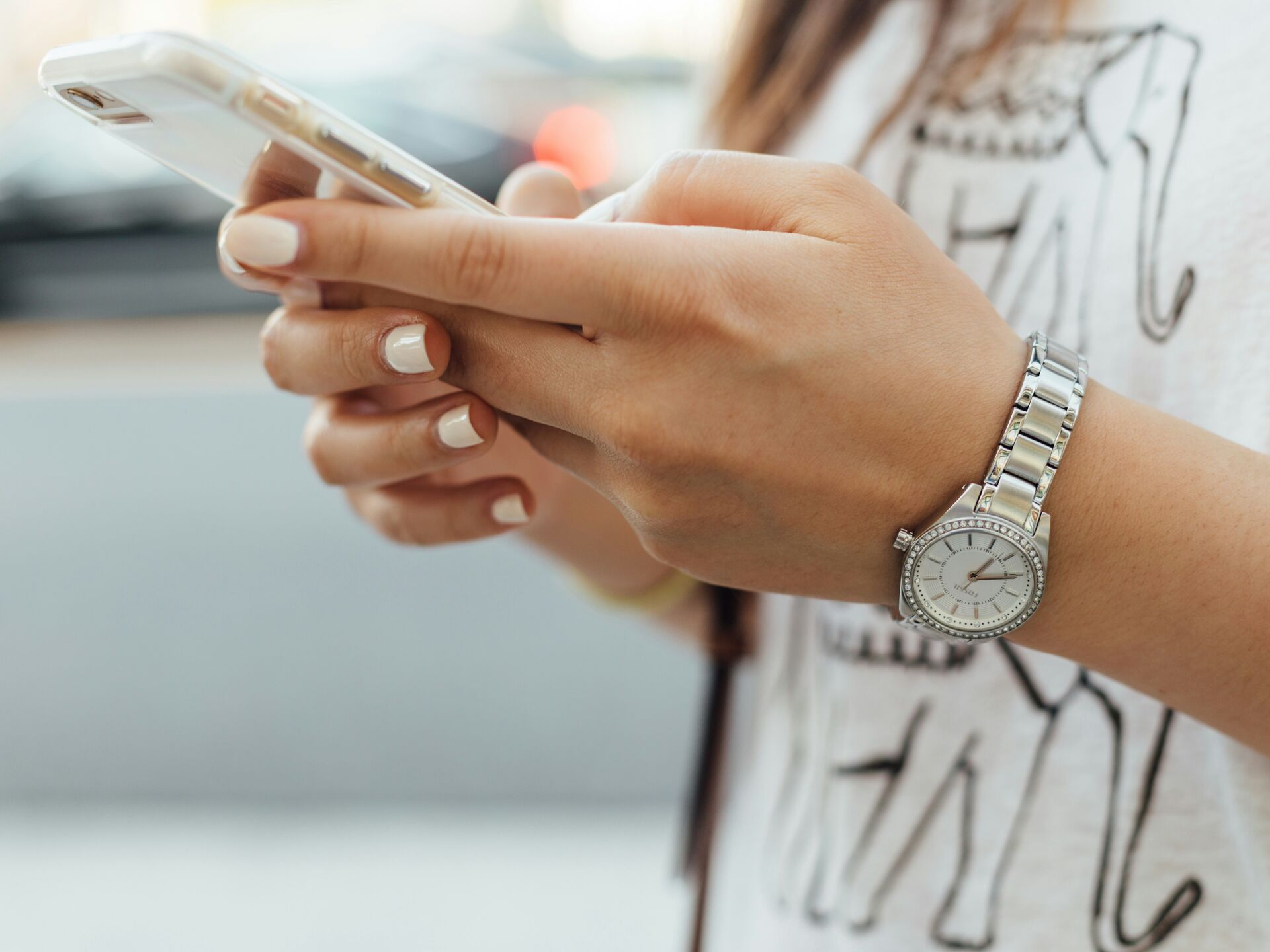 Nahaufnahme einer Person, die ein Smartphone in den Händen hält, mit silberner Armbanduhr und dezent lackierten Fingernägeln.