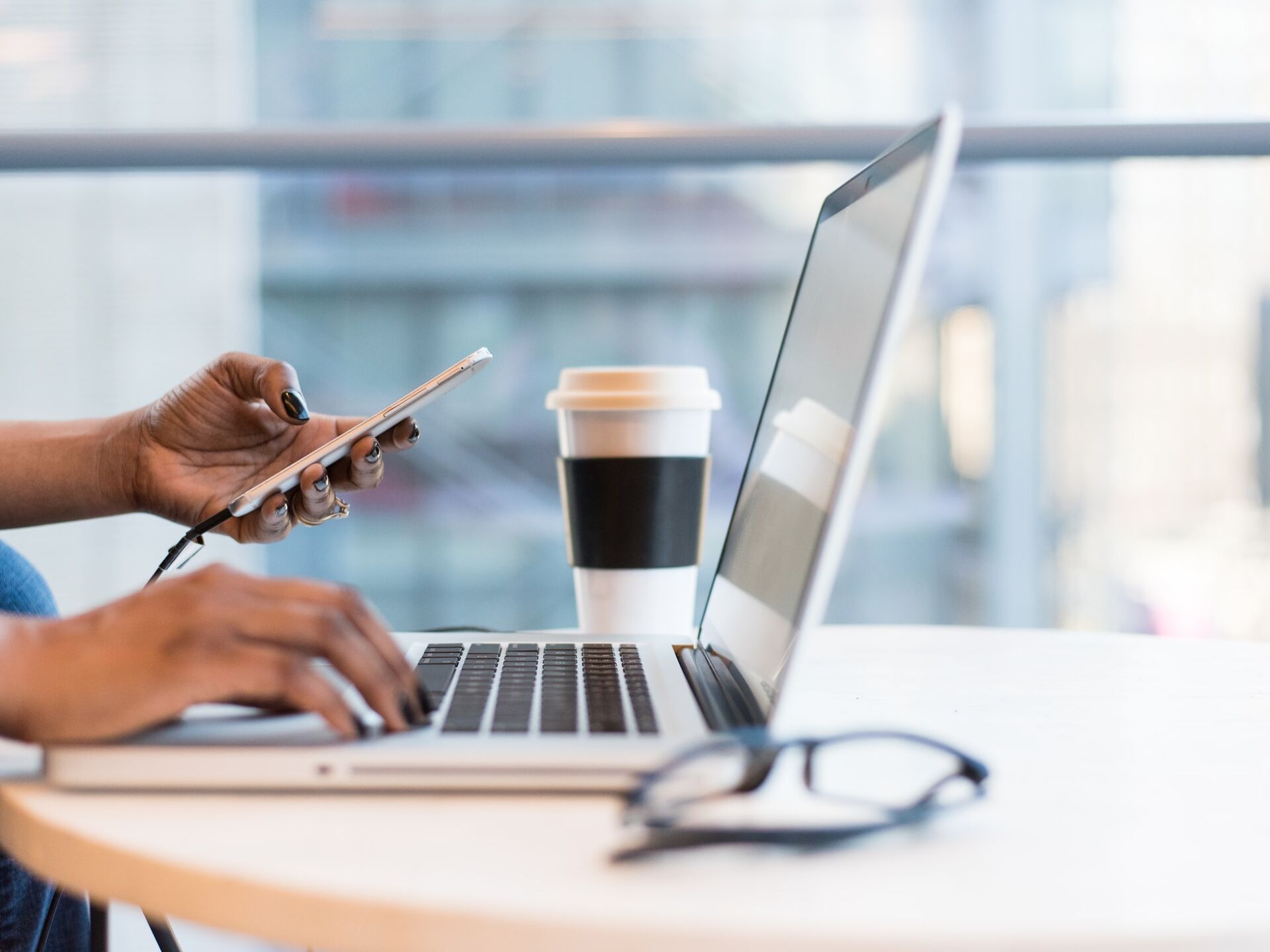 Arbeitsplatz mit Laptop, Smartphone, Kaffeebecher und Brille auf einem Tisch in modernem Umfeld.